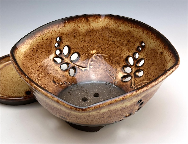 May include: A brown ceramic colander with a square shape and a decorative leaf design. The colander has small holes throughout the bottom and sides. It is sitting on a matching brown ceramic plate.