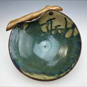 Può includere: Una ciotola in ceramica fatta a mano con un design unico. La ciotola presenta una miscela di tonalità verdi e blu, con una superficie strutturata. Un manico in legno è attaccato al lato. Il bordo della ciotola è marrone scuro. Questa ciotola decorativa è ideale per servire o esporre.