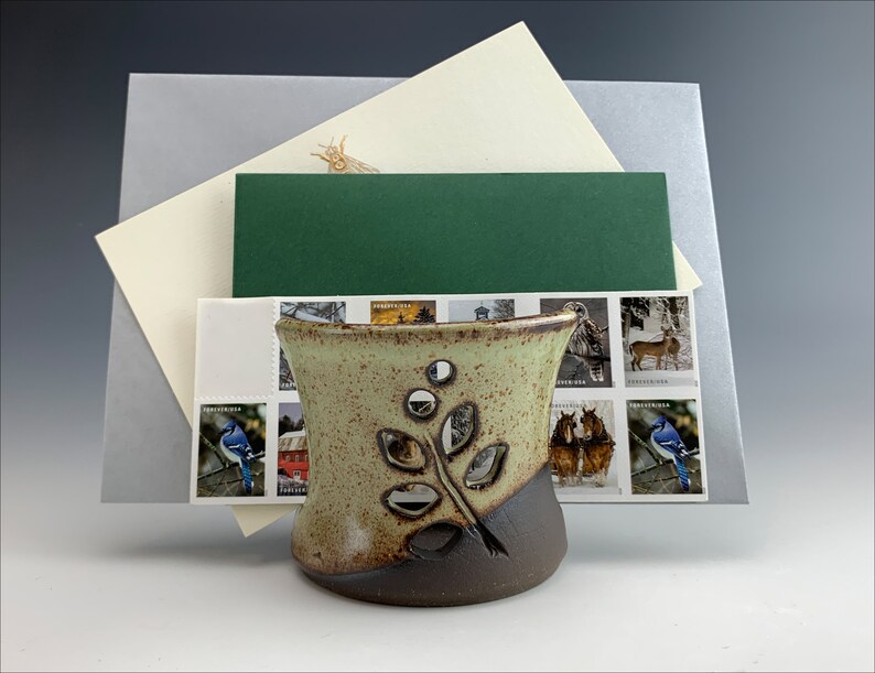 May include: A ceramic holder with a leafy design and a speckled glaze. The holder is brown and green and has a cutout design. The holder is sitting on a stack of envelopes with stamps.