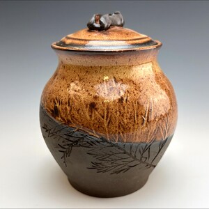Puede incluir: Un tarro de cerámica con tapa, decorado con un esmalte marrón y negro. El tarro tiene una superficie texturizada y un diseño de agujas de pino en la mitad inferior.