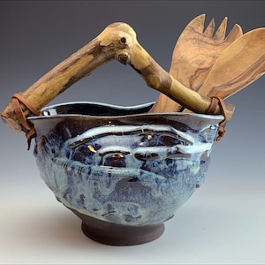 May include: A blue and brown ceramic bowl with a wooden handle and leather straps. The bowl is decorated with a blue and white glaze. Two wooden salad servers are resting in the bowl.