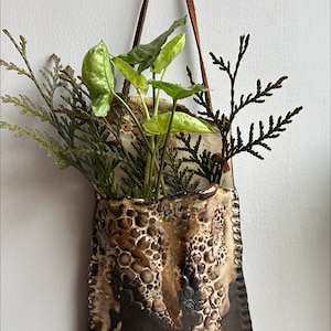 May include: A brown and white ceramic wall planter with a textured surface. The planter has a leather strap for hanging and is filled with green plants.