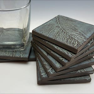 May include: A set of six ceramic coasters with a fern leaf design. The coasters are a light blue color with a dark brown border. The coasters are stacked on top of each other.
