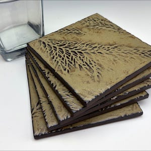 Pode incluir: Conjunto de quatro  tapetes quadrados com borda marrom e fundo marrom claro. Os tapetes apresentam um design em relevo de folhas de samambaia.