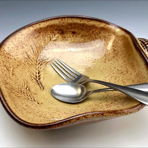 May include: A large, yellow ceramic bowl with a brown rim and a textured design of wheat stalks. The bowl is sitting on a white surface with a fork and spoon inside.