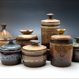 以下が含まれることがあります： 蓋付きの手作りの陶器の瓶のコレクション。瓶はサイズ、形、色が異なり、茶色、タン、青などのアースカラーが特徴です。一部はテクスチャ加工された表面と装飾的な模様があり、その他はユニークな蓋のデザインです。