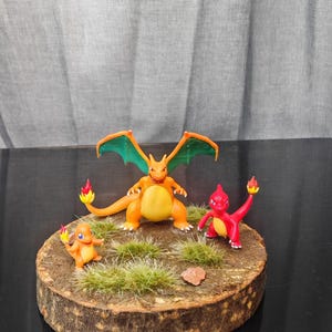 Może przedstawiać: Ręcznie wykonana diorama z trzema figurkami Pokemon: Charizard, Charmander i czerwoną postacią, wszystkie z płomieniami. Ustawione na drewnianej podstawie z zieloną trawą i małym kamieniem.