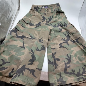 May include: Wide-leg camouflage pants in shades of green, brown, and black. The pants feature two cargo pockets on the legs and a high waist. The fabric has a classic military-inspired pattern.