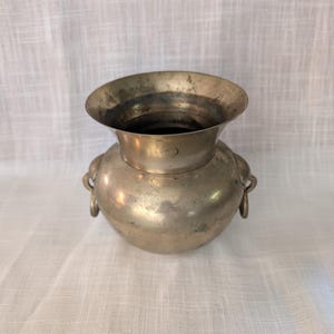 May include: A metallic, brass-toned vessel with a wide, flared rim and a rounded, bulbous body. The container features two small, circular handles attached to opposite sides of the midsection, displaying a weathered, aged patina finish.