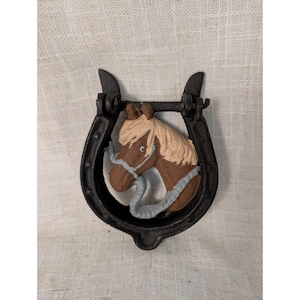 May include: A black cast iron horseshoe door knocker featuring a relief of a brown horse head with a light tan mane and a light blue bridle. The horse head is centered within the horseshoe frame, which has a hinged top section for movement.