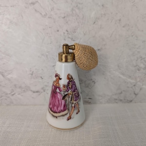 May include: A white porcelain perfume bottle with a conical shape, featuring a colorful illustration of two figures in historical attire. The bottle is topped with a gold-toned metal atomizer pump and a textured, round, gold-colored mesh bulb.