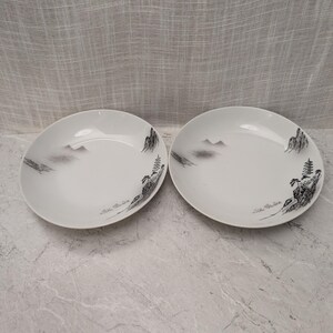 May include: Two white ceramic plates featuring a black and gray ink-wash style landscape illustration. The design depicts mountains, trees, and a pagoda structure along the rim of each plate, creating a subtle, artistic scene on the surface.