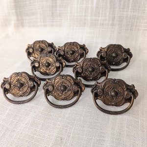 May include: A set of seven antique-style brass drawer pulls, each featuring an ornate, circular central medallion with intricate floral-inspired detailing and a matching semi-circular drop handle attached to a decorative, scalloped backplate.