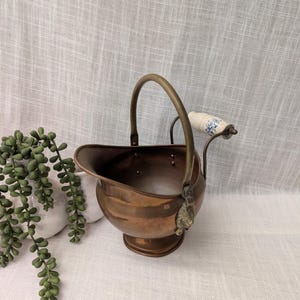 May include: A polished copper coal scuttle with a rounded body, a pedestal base, and a curved brass handle. The handle features a white ceramic grip decorated with a blue floral pattern and is attached to the vessel with ornate brass mounts.