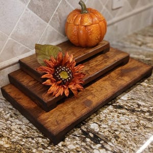 May include: A tiered wooden serving tray with a decorative orange pumpkin-shaped container and a large orange sunflower. The tray is stained a dark brown and sits on a granite countertop. The pumpkin container has a lid.