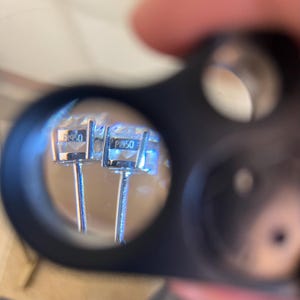 May include: A pair of silver-toned stud earrings viewed through a magnifying loupe. Each earring features a clear faceted stone set in a metal basket setting. The side of each setting is clearly stamped with the text PT950, indicating platinum.