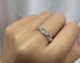 May include: A silver-toned ring featuring an irregular, organic band shape with a smooth, metallic finish. The center of the ring is set with a single, small, round, lime-green gemstone held in place by a simple prong setting.