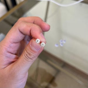 May include: A pair of round, brilliant-cut clear stud earrings set in a metallic prong setting. The earrings feature a single faceted stone each, reflecting light with a clear, sparkling appearance, held between a thumb and index finger.