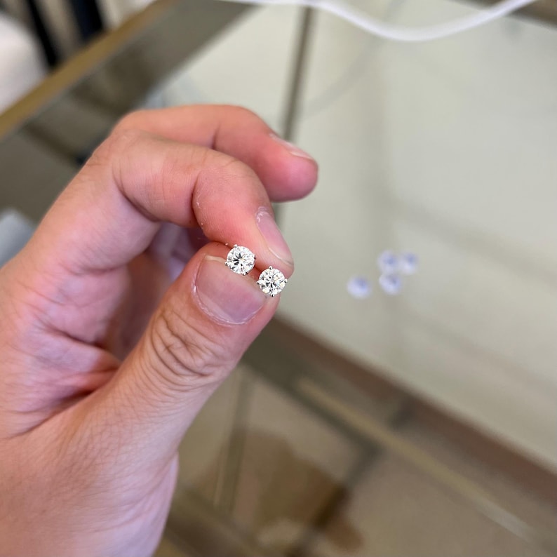 May include: A pair of round, brilliant-cut clear stud earrings set in a metallic prong setting. The earrings feature a single faceted stone each, reflecting light with a clear, sparkling appearance, held between a thumb and index finger.