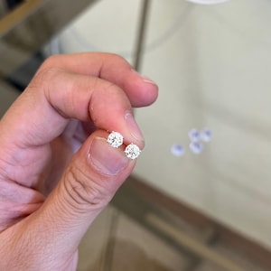 May include: A pair of round, brilliant-cut clear stud earrings set in a four-prong metal setting. The earrings feature a faceted design that reflects light, showcasing a classic circular shape held between a thumb and index finger for scale.