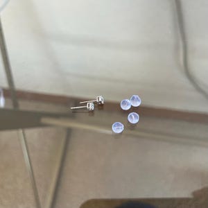 May include: Two silver-toned stud earrings featuring clear, round-cut stones in a four-prong setting, accompanied by three small, translucent, spherical silicone earring backs. The metal posts are visible extending from the back of the earring settings.