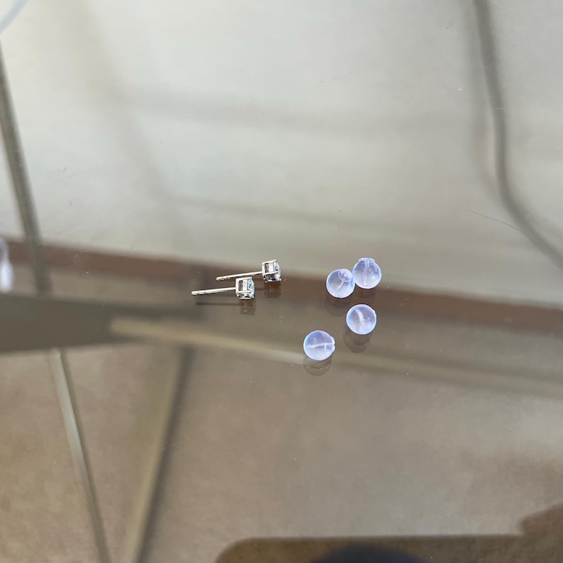 May include: Two silver-toned stud earrings featuring clear, round-cut stones in a four-prong setting, accompanied by three small, translucent, spherical silicone earring backs. The metal posts are visible extending from the back of the earring settings.