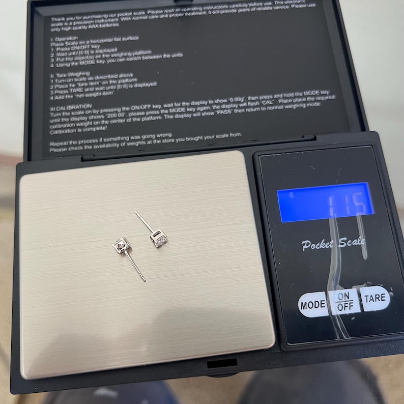 May include: Two silver-toned stud earrings with clear square-cut stones resting on a digital pocket scale. The scale display shows a weight of 1.15 grams. The scale is black with a metallic weighing platform and features buttons labeled MODE, ON/OFF, and TARE.