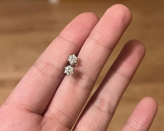 May include: A pair of round, clear gemstone stud earrings. The earrings are set in a silver metal. The gemstones are faceted, reflecting light. The earrings are small and delicate, suitable for everyday wear.