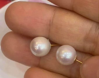 May include: A pair of round, white pearl stud earrings with gold posts. The pearls have a subtle luster and are set against a blurred background. The earrings are held in a hand.