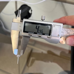 May include: A silver-toned stud earring with a clear gemstone being measured by a digital caliper. The digital display clearly shows a measurement of 4.46 mm. The earring features a central stone held by metal prongs and a thin post.
