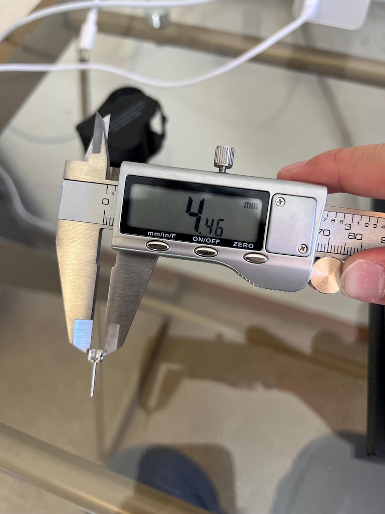 May include: A silver-toned stud earring with a clear gemstone being measured by a digital caliper. The digital display clearly shows a measurement of 4.46 mm. The earring features a central stone held by metal prongs and a thin post.