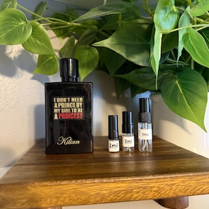 May include: A black perfume bottle with the text "I DON'T NEED A PRINCE BY MY SIDE TO BE A PRINCESS" and the brand name "Kilian". Three small sample bottles are next to the perfume bottle. The items are on a wooden tray.