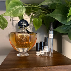May include: A perfume bottle with a dark blue cap and a black ribbon, with the word "SHALIMAR" in gold lettering. Several small sample bottles of perfume are arranged in front of the bottle, with labels indicating sizes of 1ml, 2ml, 5ml, and 10ml. The items are on a wooden tray.
