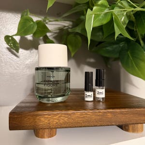 May include: A clear glass perfume bottle with a white cap and the text "FATHER FIGURE" sits on a wooden platform. Two small vials labeled "1ml" and "2ml" are next to the bottle. Green leaves are in the background.