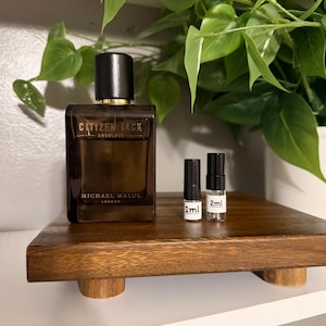 May include: A dark brown rectangular perfume bottle with a black cap and gold accents, labeled "Citizen Jack Absolute" by Michael Malul London. Two small vials labeled 1ml and 2ml are next to the bottle. The items are on a wooden tray.