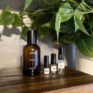 May include: A brown glass perfume bottle with a black cap, labeled "Aesop Aurher Eau de Parfum", next to three small sample vials labeled 1ml, 2ml, and 5ml. The items are on a dark wooden surface, with green leafy plants in the background.