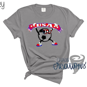 May include: Grey t-shirt with a cartoon soccer ball wearing sunglasses and holding a banner with "USA". The soccer ball has arms, legs, and is wearing red, white, and blue shoes. The word "grey" is in the top left corner.