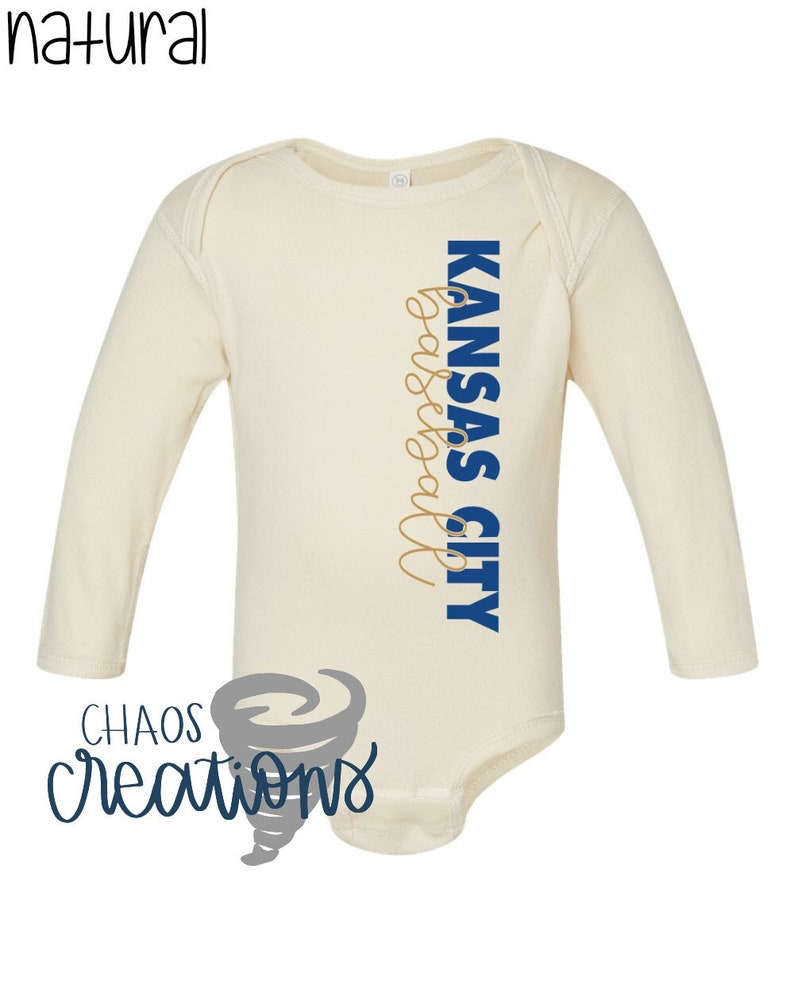 May include: A cream coloured long-sleeved baby onesie with the text "KANSAS CITY Basketball" in blue and gold lettering. The onesie has a small grey tornado design with the text "CHAOS creations" in blue lettering.