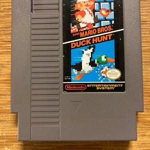 May include: A grey Nintendo Entertainment System cartridge for the game "Super Mario Bros. / Duck Hunt." The label features pixelated graphics of Mario, a duck, and a dog. The Nintendo logo and "Entertainment System" text are also visible.