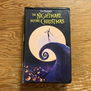 Könnte beinhalten: Eine schwarze VHS-Kassette von Tim Burton's The Nightmare Before Christmas. Das Cover zeigt eine große, dünne Figur, die auf einem geschwungenen Hügel vor einem großen gelben Mond steht, mit kleinen leuchtenden orangefarbenen Kürbissen am Hügelfuß.