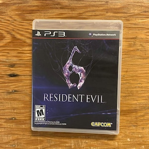 May include: A PlayStation 3 video game case for Resident Evil 6. The cover features a dark blue background with a stylized number six logo in purple and white, the Resident Evil 6 title, the PS3 logo, an ESRB M rating, and the Capcom logo.