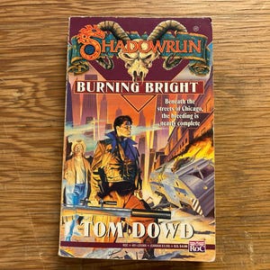 May include: A paperback book titled "Shadowrun: Burning Bright" by Tom Dowd. The cover features a futuristic cityscape with two figures and a crashed vehicle. The title is in large, bold letters.