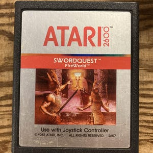 May include: Atari 2600 video game cartridge for Swordquest: FireWorld. The label features the Atari logo in red, the game title, and a fantasy illustration. The cartridge is black and has the text "Use with Joystick Controller".