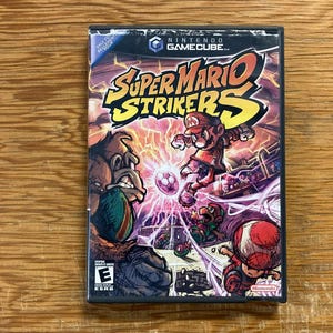 Può includere: Una custodia per videogioco Nintendo GameCube di Super Mario Strikers con un'illustrazione colorata di Mario, Donkey Kong e Toad su un campo da calcio con effetti di fulmini. Il titolo Super Mario Strikers è stampato in grandi lettere gialle.