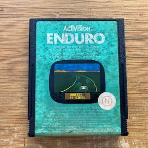 May include: A vintage Activision Enduro video game cartridge. The cartridge has a teal background with the game title in white. The game screen shows a car racing on a road. The cartridge has a black border and a round sticker with the letter "N".