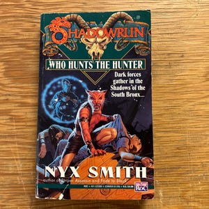 Puede incluir: Un libro de bolsillo titulado "Shadowrun: Who Hunts the Hunter" de Nyx Smith. La portada presenta un personaje humanoide pelirrojo feroz, una figura azul brillante y el texto "Dark forces gather in the Shadows of the South Bronx..."