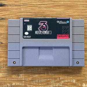 Può includere: Una cartuccia per videogiochi Super Nintendo in plastica grigia per Mortal Kombat 3. L'etichetta presenta il titolo del gioco in testo bianco, un logo del drago rosso, un logo Williams e una classificazione ESRB Mature. Ha due viti circolari.