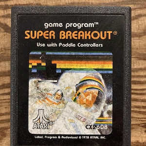 Puede incluir: Cartucho de videojuego Atari Super Breakout negro. La etiqueta presenta el título del juego en naranja, un gráfico colorido y una ilustración de un astronauta. El texto dice "Use with Paddle Controllers" y "CX2608".