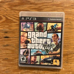 May include: A PlayStation 3 video game case for Grand Theft Auto V. The cover features a collage of various characters, vehicles, and scenes from the game, including a helicopter, a gas mask, a jet ski, and a sports car, with the M rating logo.