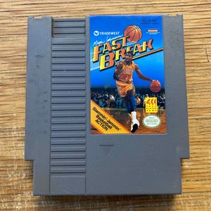 May include: A grey Nintendo Entertainment System (NES) game cartridge for the game "Fast Break." The label features an image of a basketball player in a yellow and purple uniform, with the text "Magic Johnson" and "Fast Break."